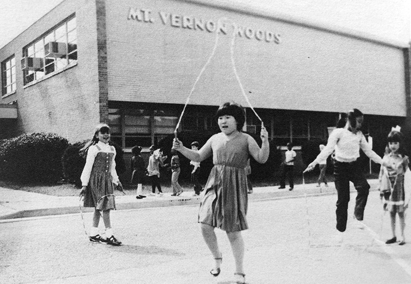 Black and white photograph of students playing on the sidewalk and in the parking lot in front of Mount Vernon Woods Elementary School. Several girls can be seen jumping rope. 